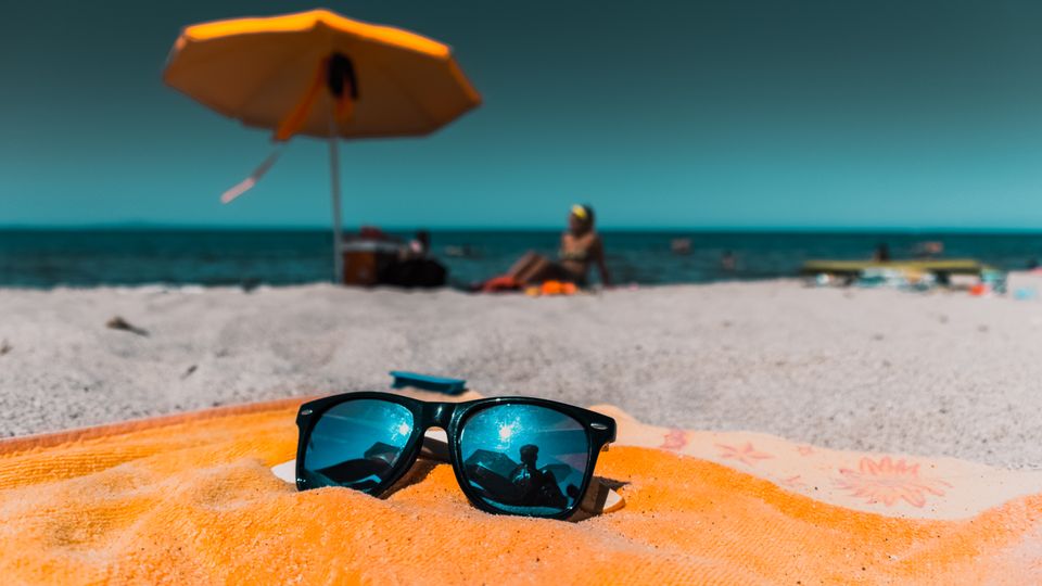 Close-up of sunglasses on an orange towel on a sunny beach with vibrant sea and umbrella.
