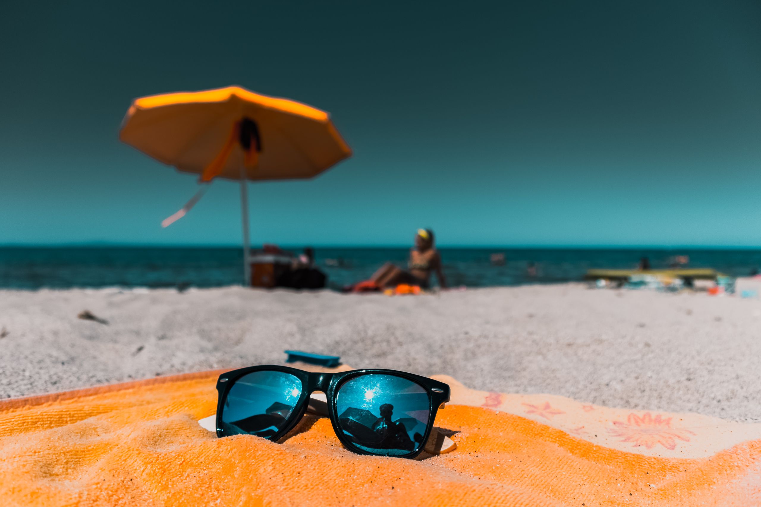 Close-up of sunglasses on an orange towel on a sunny beach with vibrant sea and umbrella.
